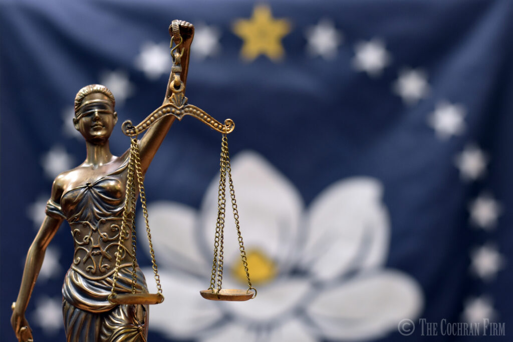 Lady Justice stands before the New Mississippi State Flag holding her sword and the Scales of Justice. 